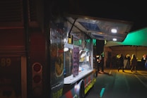 A food truck with its serving window open, illuminated by bright lights. A menu board is visible with handwritten items. In the background, there is a tent with people standing or walking, suggesting an event or gathering taking place at night.