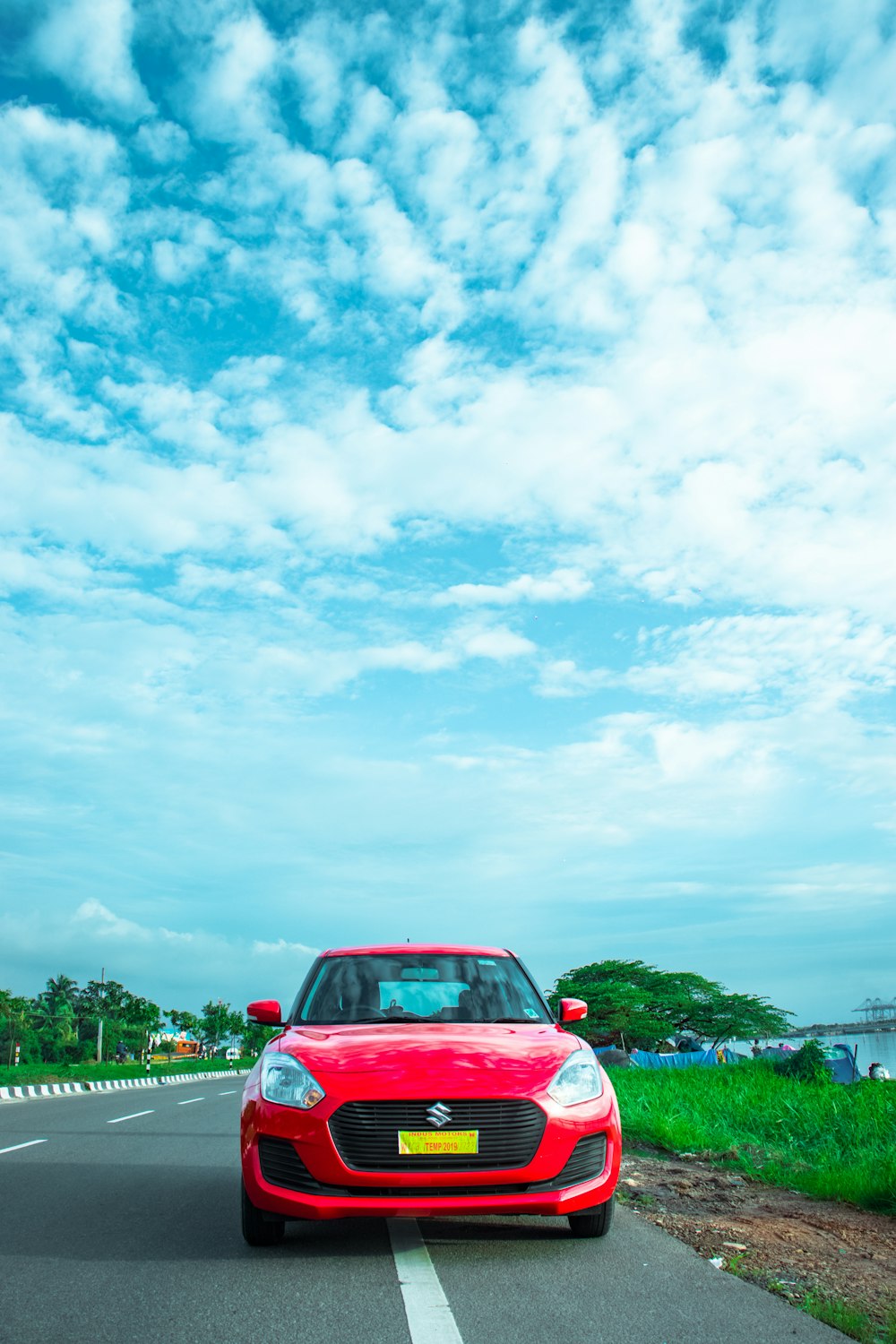 red Suzuki car on road near green field under blue and white skies during daytime