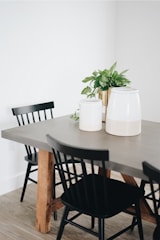 A modern dining room scene with a grey table that has wooden legs. The table is surrounded by black chairs, and on top of the table are two white canisters along with a potted green plant.