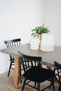 A modern dining room scene with a grey table that has wooden legs. The table is surrounded by black chairs, and on top of the table are two white canisters along with a potted green plant.