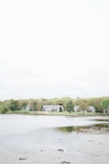 A serene landscape featuring a row of houses nestled among lush green trees by a calm body of water. The sky overhead is overcast, creating a soft and tranquil ambiance. The foreground shows a shallow expanse of water with patches of muddy ground.