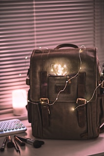 A large, brown backpack is placed on a table surrounded by a string of glowing fairy lights. Beside it, there are makeup brushes scattered near a compact keyboard, and a lit candle sits next to the bag. The background features horizontal blinds, partially open.