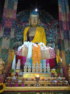 A large statue of Buddha is adorned with traditional yellow and brightly patterned garments, surrounded by colorful and intricately detailed tapestry and floral motifs. The arrangement in front of the statue includes various offerings such as bowls, flowers, and ritualistic items, with a lit candle centered among them.