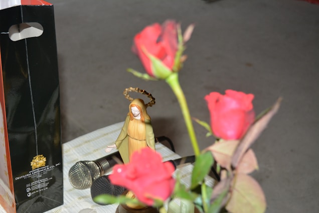 A small statuette resembling a religious figure with a golden halo is seen next to a bouquet of vibrant red roses. In the background, there is a shiny black and orange gift bag with gold lettering, and two microphones are placed on a checkered cloth surface.