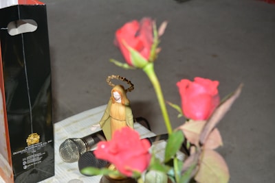 A small statuette resembling a religious figure with a golden halo is seen next to a bouquet of vibrant red roses. In the background, there is a shiny black and orange gift bag with gold lettering, and two microphones are placed on a checkered cloth surface.