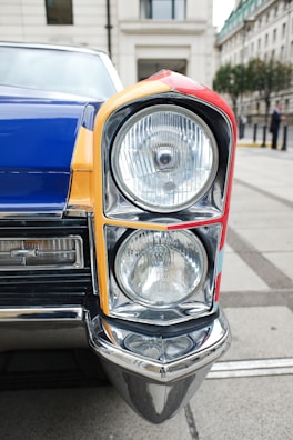 A close-up view of a vintage car's headlight and front corner. The vehicle features a colorful design with sections of red, yellow, and blue. The chrome and glass of the headlight are highly reflective, and the surrounding architecture of the street creates a classic urban backdrop.