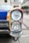 A close-up view of a vintage car's headlight and front corner. The vehicle features a colorful design with sections of red, yellow, and blue. The chrome and glass of the headlight are highly reflective, and the surrounding architecture of the street creates a classic urban backdrop.