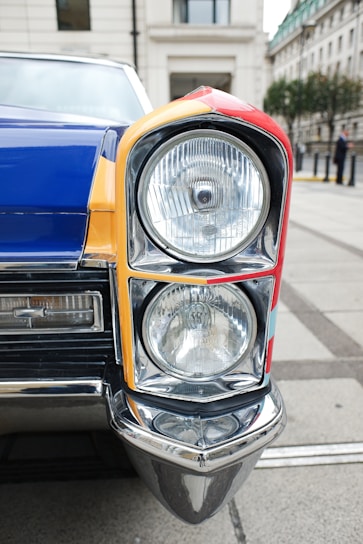 A close-up view of a vintage car's headlight and front corner. The vehicle features a colorful design with sections of red, yellow, and blue. The chrome and glass of the headlight are highly reflective, and the surrounding architecture of the street creates a classic urban backdrop.