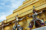 Three ornate statues, richly adorned with multicolored mosaics, stand against a backdrop of a gold structure. The statues have intricate details, featuring traditional Thai mythological figures with elaborate headpieces and body armor. The vibrant colors and patterns contrast with the gleaming gold surface, set against a clear blue sky.