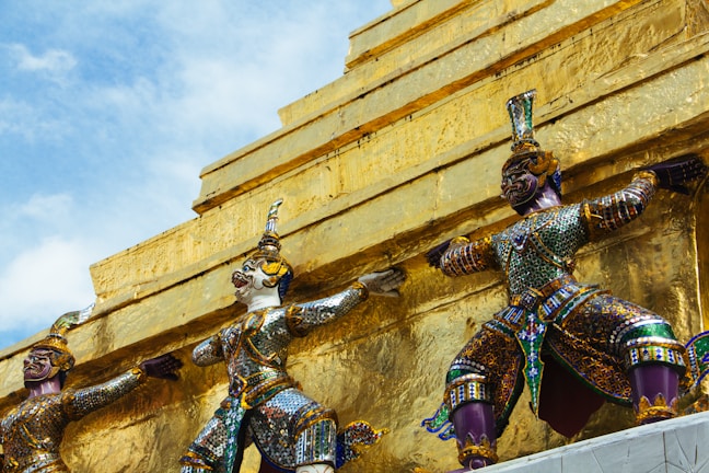 Three ornate statues, richly adorned with multicolored mosaics, stand against a backdrop of a gold structure. The statues have intricate details, featuring traditional Thai mythological figures with elaborate headpieces and body armor. The vibrant colors and patterns contrast with the gleaming gold surface, set against a clear blue sky.