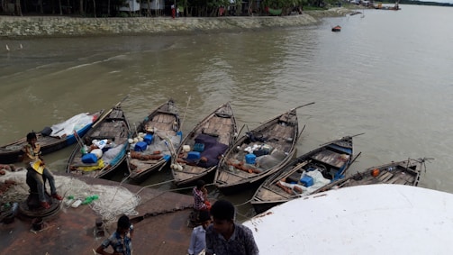 Several small wooden boats are moored alongside a riverbank. The boats contain various goods and equipment, including blue and white barrels and nets. A few people, including children, are seen near the boats and appear to be involved in fishing activities. The river looks calm, and the surrounding area is green with some structures visible in the background.