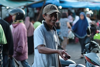 A man wearing a hat and a light-colored polo shirt stands smiling in a busy street market scene. He appears to be offering something in his hand, with a motorcycle nearby. The background is blurred with several people walking, some dressed in colorful clothing, indicating a lively atmosphere.