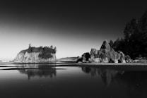 A black and white photograph capturing a serene coastal scene with a rocky shore. Large rock formations rise dramatically from the water, with a dense cluster of trees atop the distant cliffs. The calm water reflects the structures, creating a mirrored effect. The sky is clear, adding a minimalistic contrast to the rugged landscape.