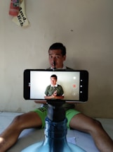A person is sitting on the floor in a relaxed position with a smartphone mounted on a water dispenser in the foreground. The phone displays the same individual, capturing a video. The setting appears simple with a plain wall and an upside-down cloth or sign in the background.