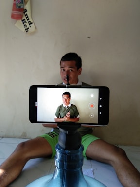 A person is sitting on the floor in a relaxed position with a smartphone mounted on a water dispenser in the foreground. The phone displays the same individual, capturing a video. The setting appears simple with a plain wall and an upside-down cloth or sign in the background.