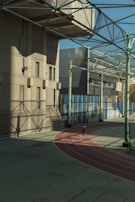 A person is running on an outdoor track surrounded by large buildings and a metal framework overhead. The track is red and curves around a large, shadowed area. The sunlight casts intricate shadows across the surface, creating a complex geometric pattern. The scene conveys a sense of urban athleticism.
