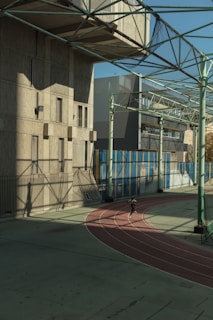A person is running on an outdoor track surrounded by large buildings and a metal framework overhead. The track is red and curves around a large, shadowed area. The sunlight casts intricate shadows across the surface, creating a complex geometric pattern. The scene conveys a sense of urban athleticism.