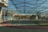 A spacious indoor sports facility with a tennis court and running tracks. The court is surrounded by high nets and tall, green metal structures supporting a roof. The area is well-lit with natural light filtering in through a transparent ceiling. In the background, there are trees with autumn foliage and several residential buildings.