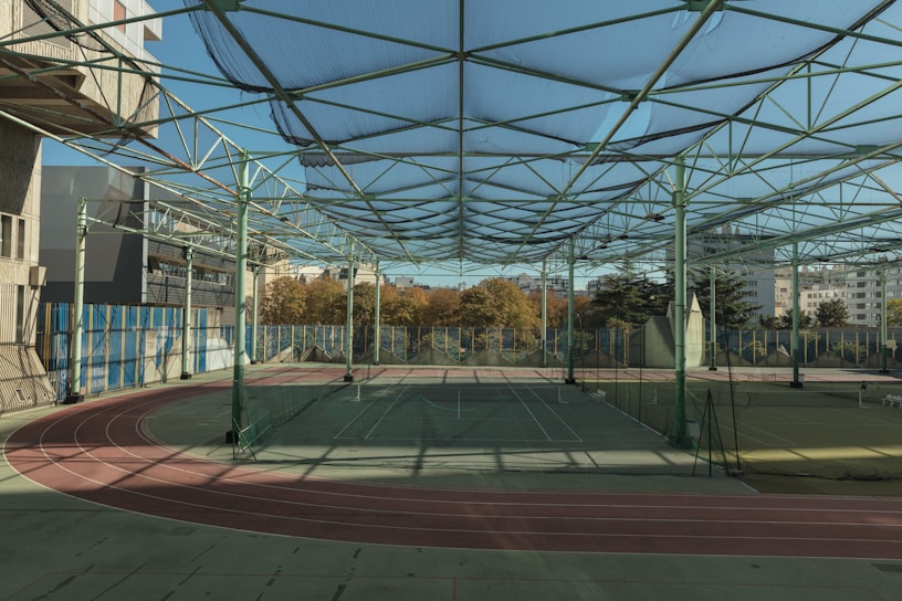A spacious indoor sports facility with a tennis court and running tracks. The court is surrounded by high nets and tall, green metal structures supporting a roof. The area is well-lit with natural light filtering in through a transparent ceiling. In the background, there are trees with autumn foliage and several residential buildings.