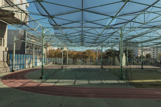 A spacious indoor sports facility with a tennis court and running tracks. The court is surrounded by high nets and tall, green metal structures supporting a roof. The area is well-lit with natural light filtering in through a transparent ceiling. In the background, there are trees with autumn foliage and several residential buildings.