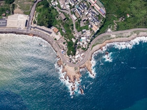 An aerial view of a coastal area featuring a blend of residential buildings and dense greenery. The coastline curves around a road leading through the area, with the sea demonstrating a gradient of blues and frothy white waves crashing against rocks. The buildings are tightly packed near the shore, and there are a few larger structures near an open parking area.