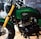 A close-up view of a custom motorcycle featuring a green fuel tank with a 'Jokowi' logo. The motorcycle has gold front forks and a prominent black engine. The environment suggests a showroom or exhibition setting with polished wooden floors and another motorcycle partially visible in the background.