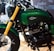 A close-up view of a custom motorcycle featuring a green fuel tank with a 'Jokowi' logo. The motorcycle has gold front forks and a prominent black engine. The environment suggests a showroom or exhibition setting with polished wooden floors and another motorcycle partially visible in the background.