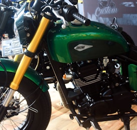 A close-up view of a custom motorcycle featuring a green fuel tank with a 'Jokowi' logo. The motorcycle has gold front forks and a prominent black engine. The environment suggests a showroom or exhibition setting with polished wooden floors and another motorcycle partially visible in the background.