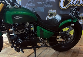 A green motorcycle with a sleek design is displayed indoors on wooden flooring. The bike features a streamlined fuel tank with the word 'Jokowi' on it, a black seat, and prominent black engine components. Behind the motorcycle, there are banners with custom design text and graphics.