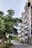 A modern apartment building with multiple balconies lines a cobblestone street. Large, leafy trees create a natural canopy along the sidewalk, offering a contrast between urban and natural elements. The view includes a clear sky and a few parked vehicles in the background.