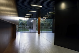A modern interior space with a sleek design featuring tiled flooring and wood paneling. Large glass doors at the end of the corridor offer a view of a neatly maintained garden with green foliage. The ceiling has inset lighting that casts a soft glow.