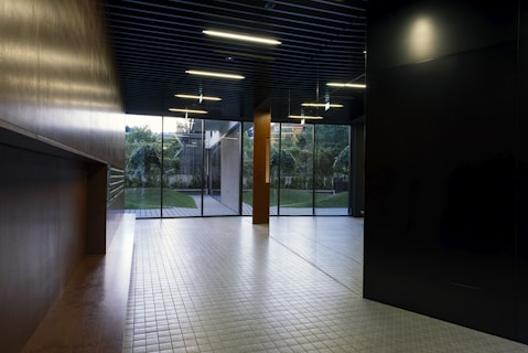 A modern interior space with a sleek design featuring tiled flooring and wood paneling. Large glass doors at the end of the corridor offer a view of a neatly maintained garden with green foliage. The ceiling has inset lighting that casts a soft glow.