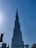 A tall, iconic skyscraper dominates the skyline against a clear blue sky. The structure has a futuristic design with multiple tiers and appears very modern and sleek. Several smaller buildings and construction cranes are visible at the base, highlighting urban development.