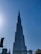 A tall, iconic skyscraper dominates the skyline against a clear blue sky. The structure has a futuristic design with multiple tiers and appears very modern and sleek. Several smaller buildings and construction cranes are visible at the base, highlighting urban development.