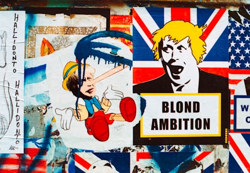 A collage of street art featuring satirical political imagery. The central focus includes a depiction of a child-like character with an elongated nose and blue paint dripping over the face, juxtaposed with another stylized image of a person's face with yellow hair above a sign that reads 'BLOND AMBITION'. The background includes various elements resembling the Union Jack flag.