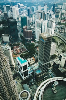 A panoramic view of a dense urban cityscape featuring numerous high-rise buildings, skyscrapers, and modern architecture. The layout of the buildings suggests a bustling metropolitan area with a mix of glass and steel structures. A swimming pool can be seen atop one of the buildings, indicating luxury amenities. The foreground includes the architectural details of the Petronas Towers.