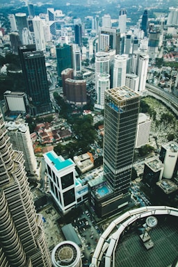 A panoramic view of a dense urban cityscape featuring numerous high-rise buildings, skyscrapers, and modern architecture. The layout of the buildings suggests a bustling metropolitan area with a mix of glass and steel structures. A swimming pool can be seen atop one of the buildings, indicating luxury amenities. The foreground includes the architectural details of the Petronas Towers.