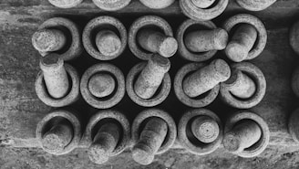 A black and white arrangement of multiple stone mortars and pestles aligned in an organized pattern. Each mortar holds one pestle, showcasing a circular and cylindrical design. The texture of the stone is rough and rustic.