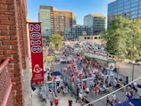 A vibrant outdoor scene featuring a large crowd of people gathered near a sports venue. A prominent red banner with '2018' and a sports logo hangs from a brick building. Nearby, there is a sign for a team store and souvenir shop. Surrounding the event are several modern high-rise buildings and a parking lot full of cars. Tall trees are visible in the background.