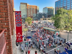 A vibrant outdoor scene featuring a large crowd of people gathered near a sports venue. A prominent red banner with '2018' and a sports logo hangs from a brick building. Nearby, there is a sign for a team store and souvenir shop. Surrounding the event are several modern high-rise buildings and a parking lot full of cars. Tall trees are visible in the background.