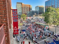 A vibrant outdoor scene featuring a large crowd of people gathered near a sports venue. A prominent red banner with '2018' and a sports logo hangs from a brick building. Nearby, there is a sign for a team store and souvenir shop. Surrounding the event are several modern high-rise buildings and a parking lot full of cars. Tall trees are visible in the background.