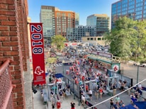 A vibrant outdoor scene featuring a large crowd of people gathered near a sports venue. A prominent red banner with '2018' and a sports logo hangs from a brick building. Nearby, there is a sign for a team store and souvenir shop. Surrounding the event are several modern high-rise buildings and a parking lot full of cars. Tall trees are visible in the background.