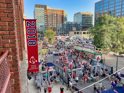 A vibrant outdoor scene featuring a large crowd of people gathered near a sports venue. A prominent red banner with '2018' and a sports logo hangs from a brick building. Nearby, there is a sign for a team store and souvenir shop. Surrounding the event are several modern high-rise buildings and a parking lot full of cars. Tall trees are visible in the background.