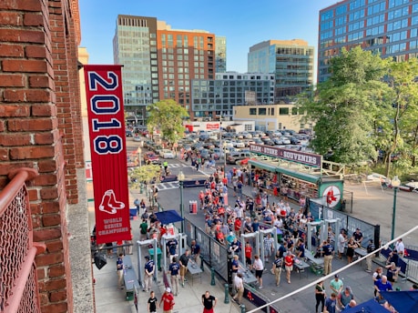 A vibrant outdoor scene featuring a large crowd of people gathered near a sports venue. A prominent red banner with '2018' and a sports logo hangs from a brick building. Nearby, there is a sign for a team store and souvenir shop. Surrounding the event are several modern high-rise buildings and a parking lot full of cars. Tall trees are visible in the background.