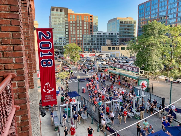 A vibrant outdoor scene featuring a large crowd of people gathered near a sports venue. A prominent red banner with '2018' and a sports logo hangs from a brick building. Nearby, there is a sign for a team store and souvenir shop. Surrounding the event are several modern high-rise buildings and a parking lot full of cars. Tall trees are visible in the background.