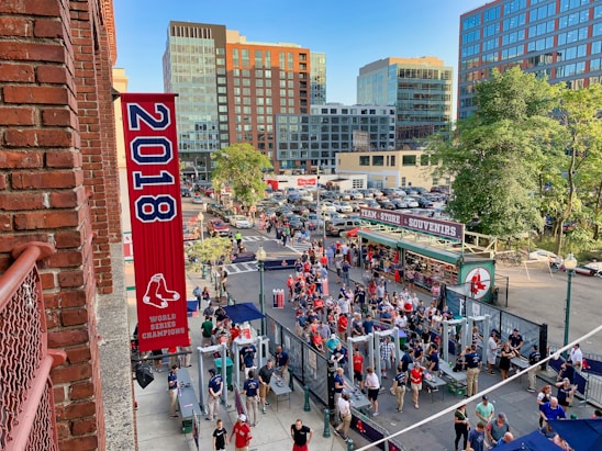 A vibrant outdoor scene featuring a large crowd of people gathered near a sports venue. A prominent red banner with '2018' and a sports logo hangs from a brick building. Nearby, there is a sign for a team store and souvenir shop. Surrounding the event are several modern high-rise buildings and a parking lot full of cars. Tall trees are visible in the background.
