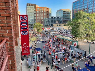 A vibrant outdoor scene featuring a large crowd of people gathered near a sports venue. A prominent red banner with '2018' and a sports logo hangs from a brick building. Nearby, there is a sign for a team store and souvenir shop. Surrounding the event are several modern high-rise buildings and a parking lot full of cars. Tall trees are visible in the background.