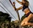 A female athlete is captured in mid-air action as she jumps near a volleyball net. She wears a sports bra and shorts, with tattoos visible on her arm and is surrounded by a backdrop of greenery and palm trees.