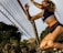 A female athlete is captured in mid-air action as she jumps near a volleyball net. She wears a sports bra and shorts, with tattoos visible on her arm and is surrounded by a backdrop of greenery and palm trees.