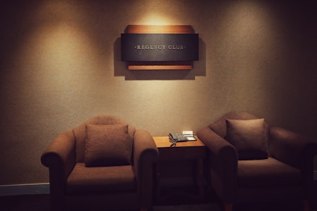 A cozy and dimly lit room features two comfortable brown armchairs positioned on either side of a small wooden table. Mounted on the wall above the table is a decorative sign that reads 'Regency Club.' The table holds a classic telephone and some note pads.